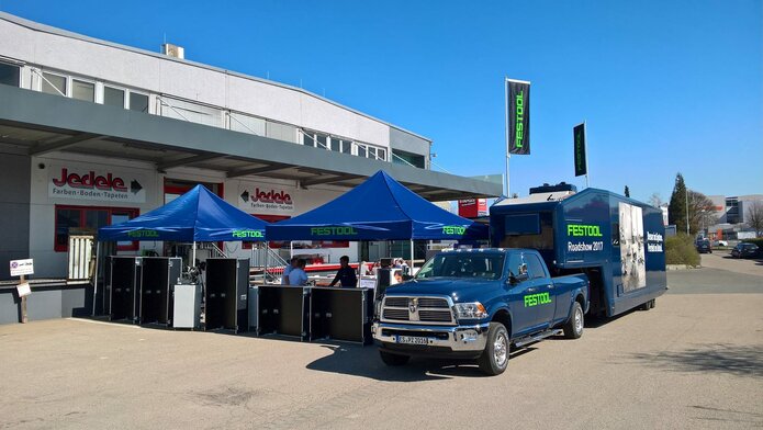 3x3m blaues Eventzelt mit grünem Festool-Logo kombiniert mit Firmenwagen und Wohnwagen vor dem Baumarkt Jedele