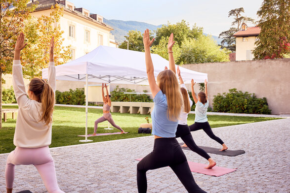 Der weiße Faltpavillon ist bei einem Yogakurs im Einsatz. Die Yoga-Leiterin steht darunter. Davor sind die Teilnehmer am Yogakurs.