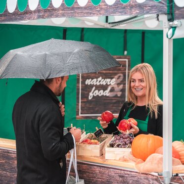 Die Verkäuferin im Marktzelt hält verschiedenes Obst in der Hand. Der Kunde steht vor dem Verkaufszelt an der Theke.