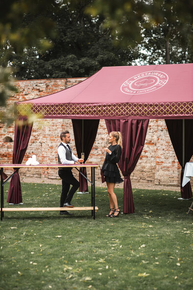 Il gazebo catering, di colore bordeaux, si trova nell'ampio giardino. La donna e l'uomo hanno un bicchiere di champagne in mano e sono in piedi davanti ad un tavolo alto.