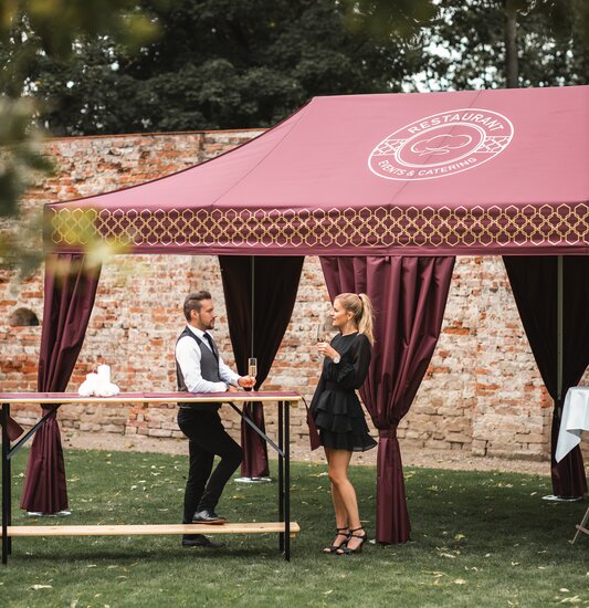 Il gazebo catering, di colore bordeaux, si trova nell'ampio giardino. La donna e l'uomo hanno un bicchiere di champagne in mano e sono in piedi davanti ad un tavolo alto.