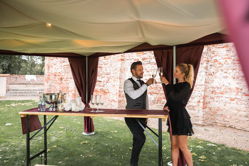 Sotto l'elegante gazebo pieghevole con cielo, 2 persone stanno in piedi con un bicchiere di prosecco in mano. 