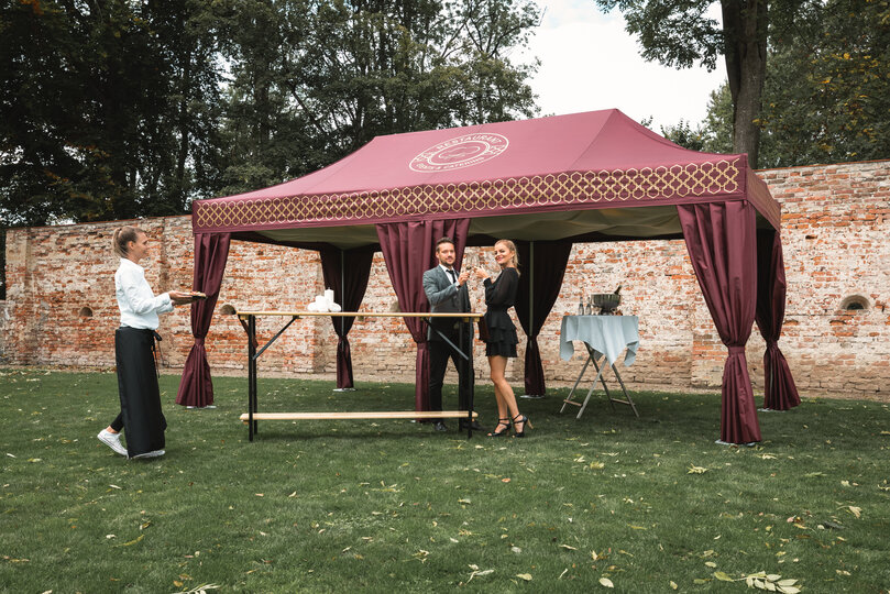 Il gazebo catering color bordeaux si trova in un prato. Una coppia è in piedi davanti ad essa con un bicchiere di prosecco in mano. La cameriera porta degli stuzzichini su un vassoio.