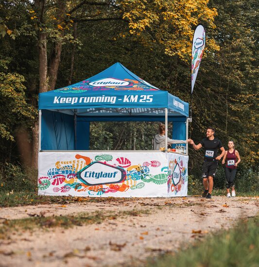 Gazebo per eventi blu 3x3m con stampa personalizzata Citylauf, con bandiera personalizzata, parete laterale chiusa e parete laterale a mezza altezza con bancone utilizzato come punto di ristoro a una corsa cittadina